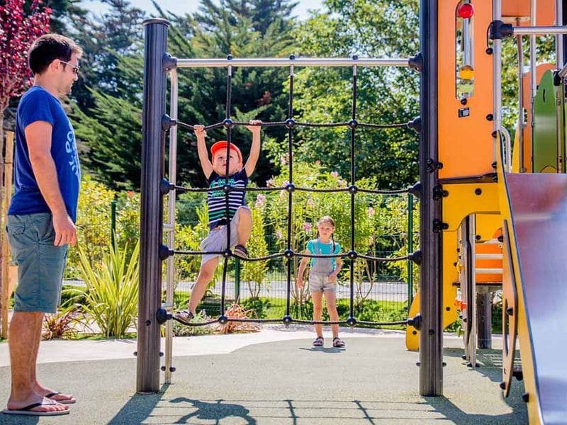 Little boy with cap on a climbing frame with slide. Father stands watching, little girl waits in the background.