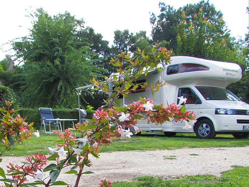 Camping La Poterie: motorhome and folding chairs with table. Branches with pink and white flowers in the foreground.