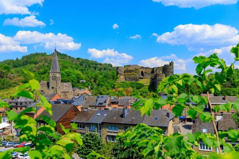 Uitzicht over La Roche-en-Ardenne met de kerktoren, de historische kasteelruïne op een heuvel en de groene Ardense natuur onder een blauwe lucht met witte wolken.