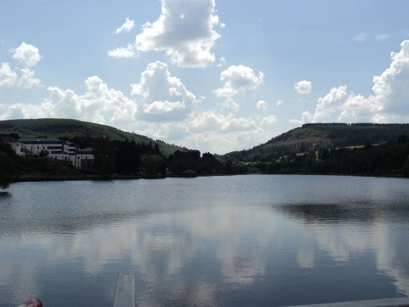Lac des Doyards in Vielsalm, een rustig meer in de Belgische Ardennen, omringd door beboste heuvels en groene natuur onder een blauwe lucht met wolken.